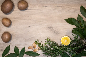 Light wooden board framed with fresh rosemary sprigs, bay leaves, lemon half, cloves of garlic and few raw potatoes, design template 
