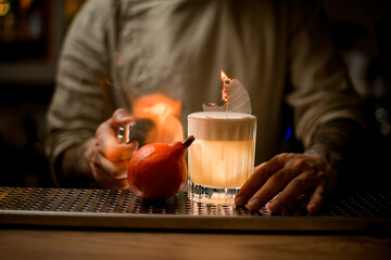 bartender splashes and sets fire to glass of cocktail with foam and decorative leaf and pumpkin on bar.