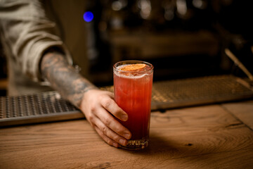 barman's hand holds glass with cold drink with ice cubes, decorate with orange slice