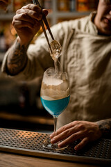 male barman gently holds piece of ice over steaming wine glass decorated with gold