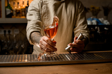 Male bartender holds jigger and cocktail glass with splashing alcoholic drink in his hands