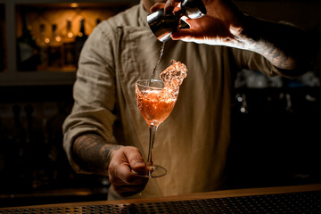Male bartender holds cocktail glass with splashing alcoholic drink and adds liquid from jigger
