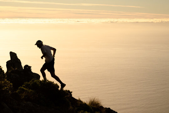 A runner silhouetted against the golden light of sunset over the ocean while running up a steep mountain trail.