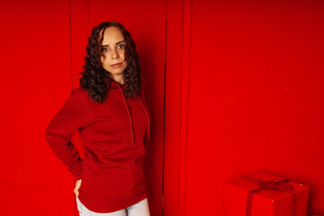 Young woman in hoodie on red background. Curly brunette poses near red wall.