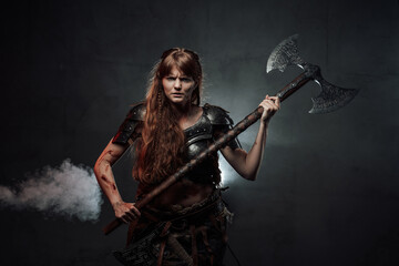 Violent and savage scandinavian female warrior holding huge two handed axe in dark foggy background.