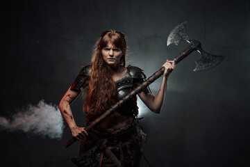 Violent and savage scandinavian female warrior holding huge two handed axe in dark foggy background.