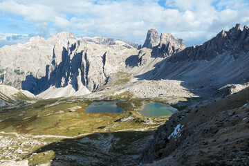 Obraz premium Small, navy blue lakes at the bottom of the valley in Italian Alps. The lakes are surrounded by high and steep peaks. The slopes are lush green. The sky is full of soft clouds. Raw landscape. Remedy