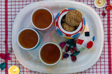 Elevated shot of tea cups and cookies pot arranged on a serving tray, placed on a red checkered table cloth, decorated with tea lights.