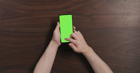 Top view man hands hold smartphone with green screen over black walnut wood table