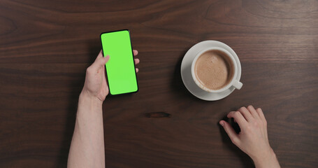 Top view man drinking hot chocolate and using smartphone with green screen over black walnut wood table