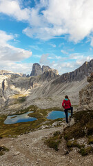 Woman hiking with the view on small, navy blue lakes at the bottom of the valley in Italian Alps. The lakes are surrounded by high and steep peaks The sky is full of soft clouds. Raw landscape. Remedy