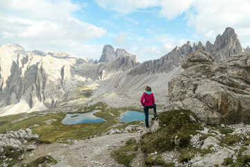 Fototapeta premium Woman hiking with the view on small, navy blue lakes at the bottom of the valley in Italian Alps. The lakes are surrounded by high and steep peaks The sky is full of soft clouds. Raw landscape. Remedy
