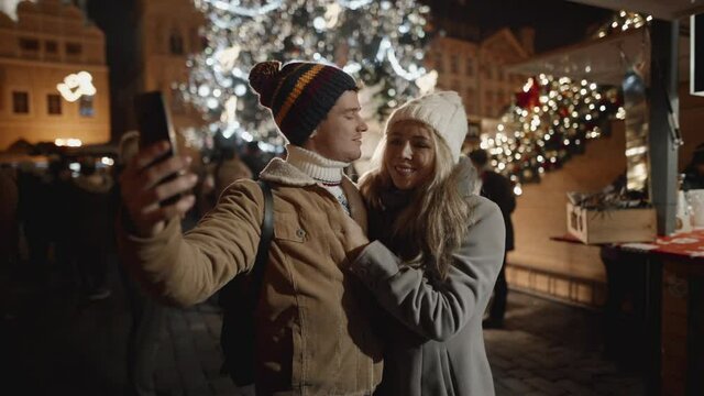 Cute scene of pretty young couple kissing while taking selfie outside on winter holiday season. Couple happy together, travelling around christmas time.