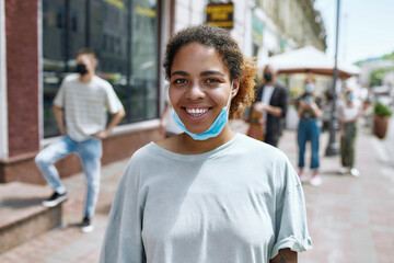 Portrait of cheerful young african american woman with mask on her chin smiling at camera. People collecting their orders from the pickup point during coronavirus lockdown in the background