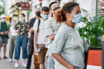Young african american woman wearing mask waiting, standing in line with other people, respecting...