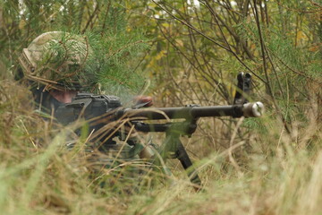 A soldier with a machine gun hid in the forest and shoots.