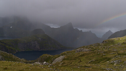 Panoramic view of Krokvatnet lake surrounded by rugged mountains near Munkebu hut on Moskenesøy island, Lofoten, Norway on stormy day in summer with rain, clouds and slight rainbow.
