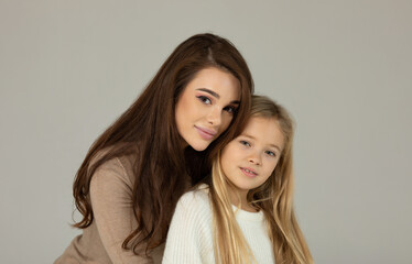 Charming little girl and her beautiful young mom are hugging looking at camera and smiling on grey background. Happy family.