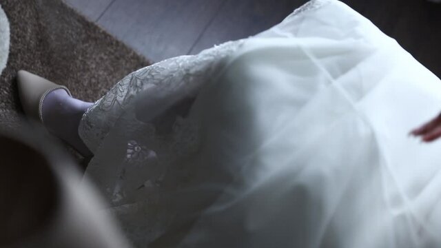 a girl bride in a white dress puts shoes on her foot in white stockings and fastens the strap on them