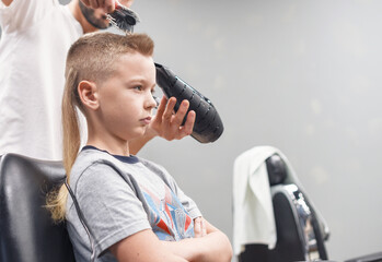 the process of hair styling a blond boy with with hair dryer and comb in a chair in a barbershop salon, a barbershop concept for men and boys