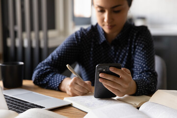 Busy indian female student write up information from phone screen to paper notebook. Young hindu lady engaged in search data online using cell take notes to textbook by hand. Focus on gadget. Close up