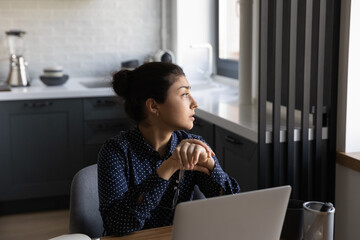 Relaxing at home office. Pensive indian female freelancer sit by work desk in kitchen look at...