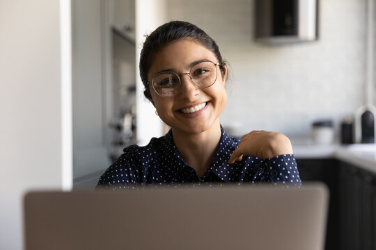 Working at home office. Portrait of happy smiling young indian female in stylish glasses sitting by laptop. Confident pretty mixed race businesswoman looking at camera posing indoor behind pc screen
