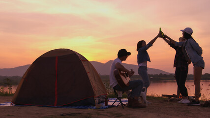 Group of four friends hanging out together at camp site playing guitar, singing song and dancing during beautiful hour of sunset