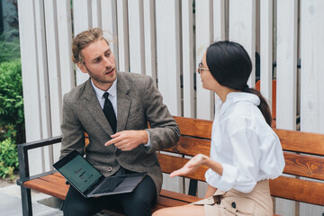 asian girl and man in business clothes on a bench with a laptop are discussing
