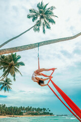 woman aerialist in white swimsuit makes aerial trick upside down on the red airsilk on the palm...