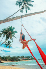 girl aerialist in white swimsuit makes aerial trick  on the red airsilk on the palm tree on the sky...