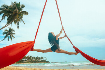 woman aerialist in black swimsuit makes aerial trick  on the red airsilk on the palm tree on the...