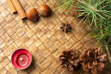 Aerial view of braided surface with red candle, pine cones, hazelnuts, cinnamon and green branches, selective focus, horizontally, with copy space