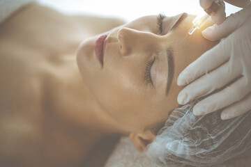 Beautician doing beauty procedure with syringe to face of young brunette woman in sunny clinic. Cosmetic medicine and surgery, beauty injections