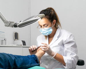 Young podiatrist doing chiropody in her podiatry clinic. The chiropodist is cutting the patient's nails with specialized scissors - obrazy, fototapety, plakaty