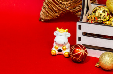 White toy bull of white color on a box background with Christmas balls of red and gold on a red background, New Year's card, place for text. Chinese Year of the Ox, symbol of 2021.