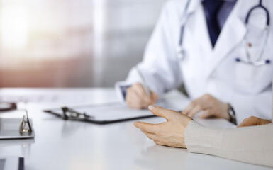 Fototapeta premium Unknown male doctor and patient woman discussing something while sittingin a darkened clinic, glare of light on the background. Close-up of hands
