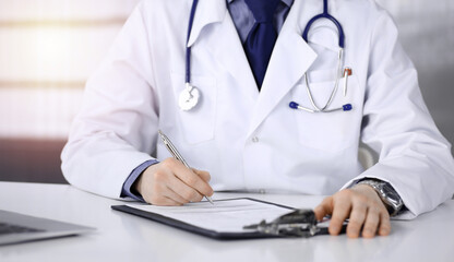 Unknown male doctor sitting and working with clipboard of medication history record in a darkened clinic, glare of light on the background, close-up of hands