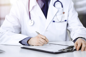 Unknown male doctor sitting and working with clipboard of medication history record in a darkened clinic, glare of light on the background, close-up of hands