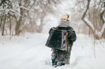 Little cheerful girl in oversized padded jacket standing on snowy road in winter day with accordion