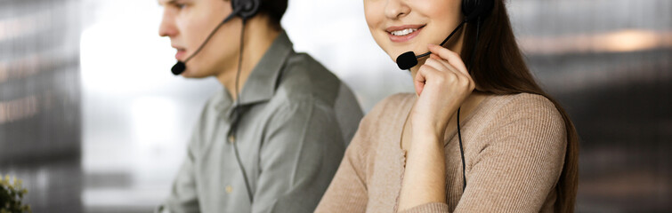 Two young people in headsets are talking to the clients, while sitting at the desk in an office....
