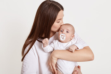Mom with newborn baby posing against light background, mommy kissing her infant with nipple, attractive female feels happy, expressing gentle, wearing white clothing.