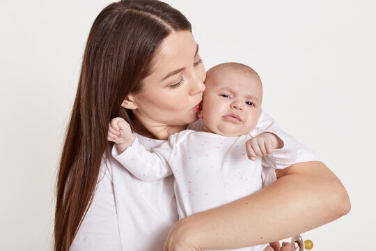Mother kissing and hugging newborn daughter isolated over white background, tender, care, lady with brown hair expressing love to her child.