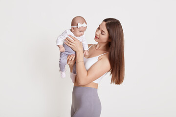 Cheerful beautiful young woman in bra and grey leggins, holding baby girl in her hands and looking at her, brown haired female posing with child against white wall.