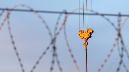 The crane's load hook is illuminated by orange rays of the sun against the sky. In the foreground is a blurred barbed wire.