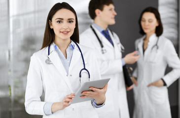 Smiling intelligent woman-doctor is holding a tablet computer in her hands, while she is standing together with her colleagues in a clinic. Physicians at work. Perfect medical service in a hospital