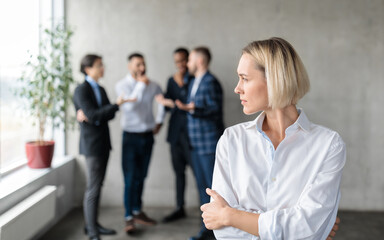 Male Coworkers Whispering Behind Back Of Unhappy Businesswoman In Office - obrazy, fototapety, plakaty