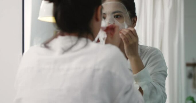 happy attractive young asian woman doing a facial mask before working in the morning look in mirror careful to smooth the mask ,close up portrait