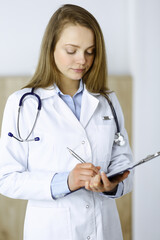 Serious female doctor using clipboard while standing in clinic. Medicine, Coronavirus pandemic concepts