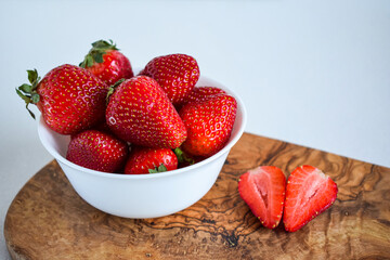 White cup with strawberries stands on a cutting wooden board. Another strawberry cut in the shape of a heart lies on a cutting board. Dessert for Valentine's Day.